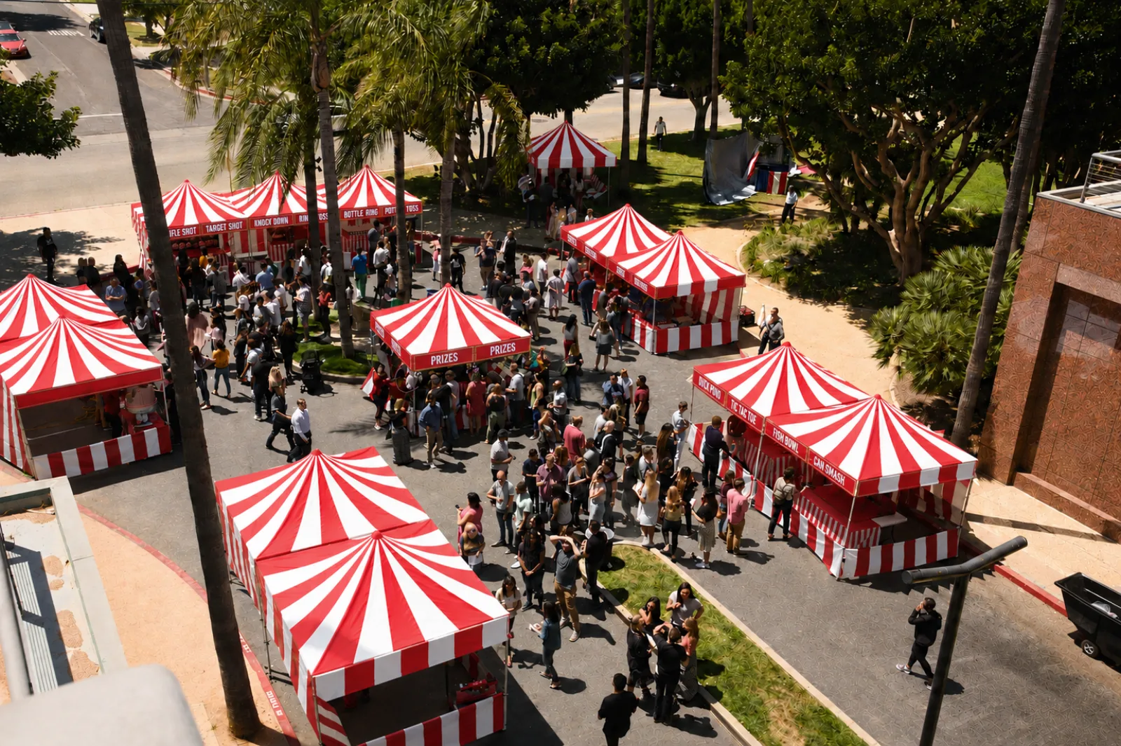 Overhead view of a dozen red-and-white striped game booths, each labeled with its game, filling a palm-lined plaza with a large crowd