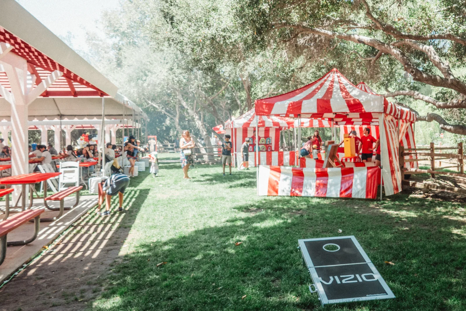 Red-and-white striped game booths under oak trees at a company family day in a park, with families at picnic tables and a branded cornhole board on the grass
