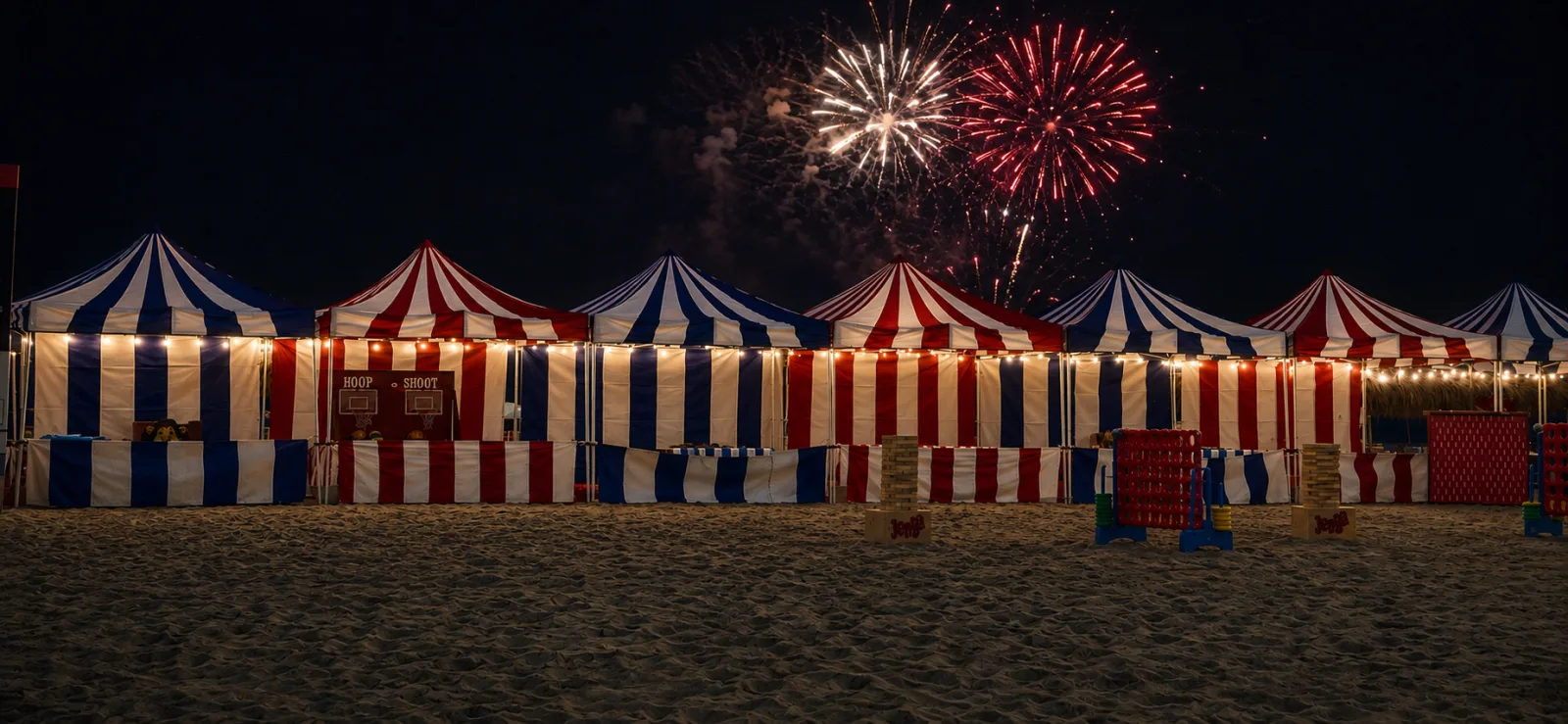 A row of red-and-white and blue-and-white striped carnival booths strung with lights on a beach at night, with fireworks bursting overhead and giant lawn games set up on the sand