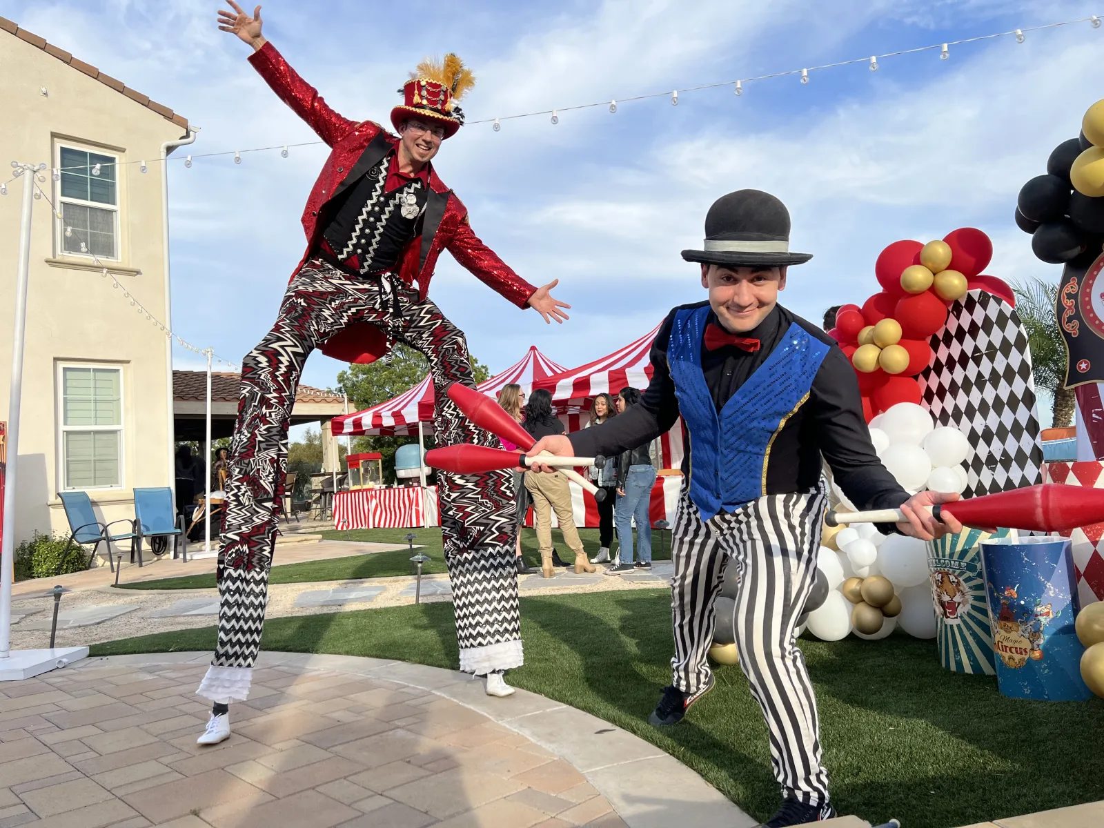 A stilt walker in a red sequined tailcoat and a juggler in a bowler hat performing beside red-and-white striped booths and balloon columns at an outdoor event