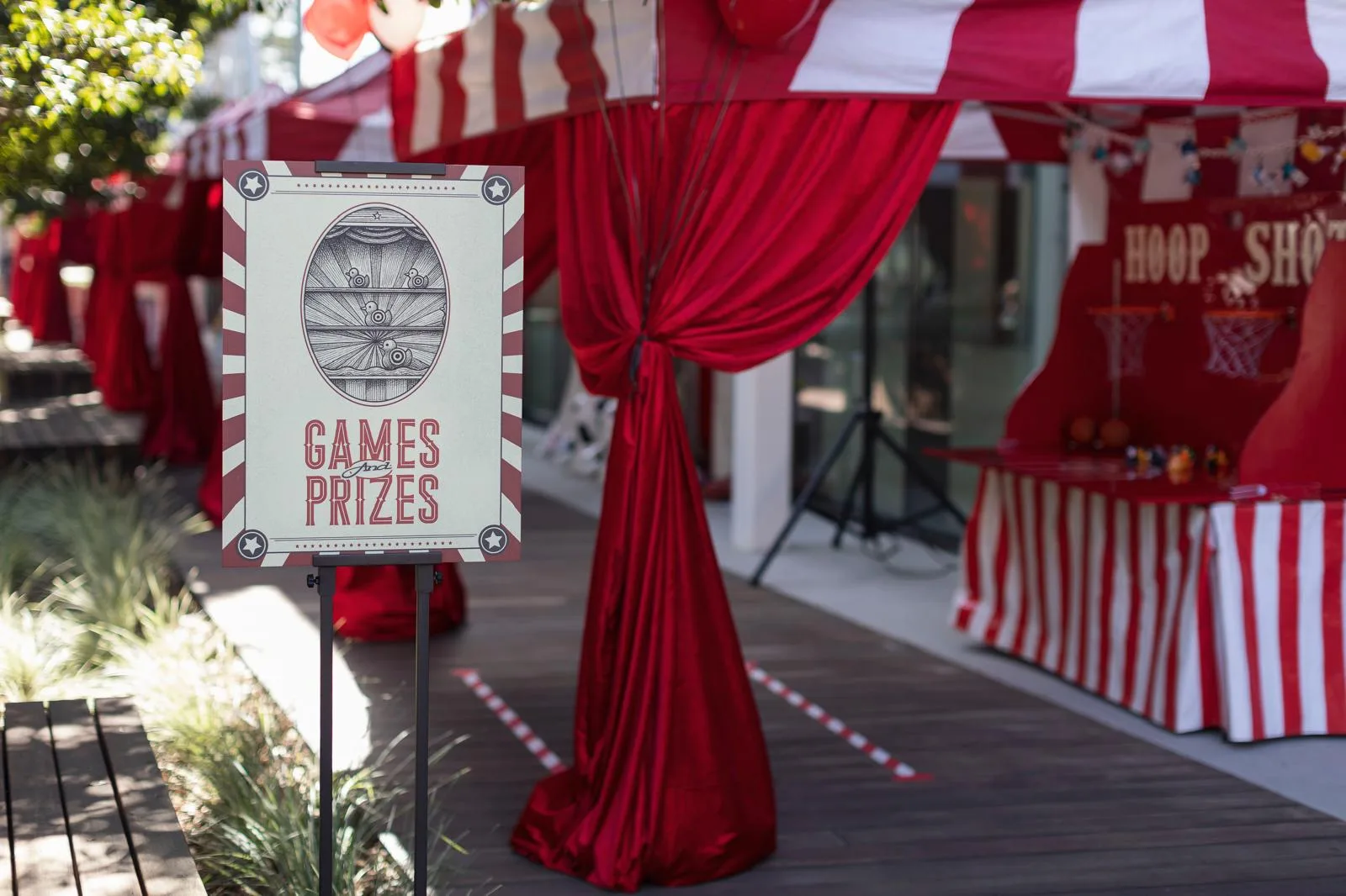 A vintage-styled Games and Prizes easel sign beside a tied-back red velvet drape and a red-and-white striped hoop-shot booth on a wooden deck