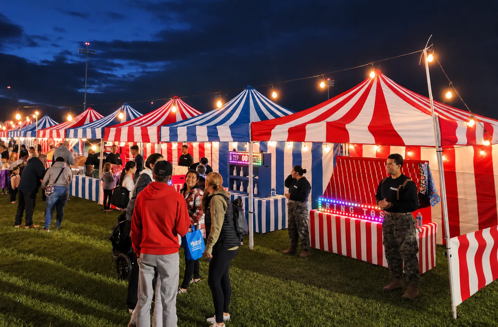 Red-and-white and blue-and-white striped carnival booths under string lights at dusk, with service members in uniform and a lit ring-toss marquee