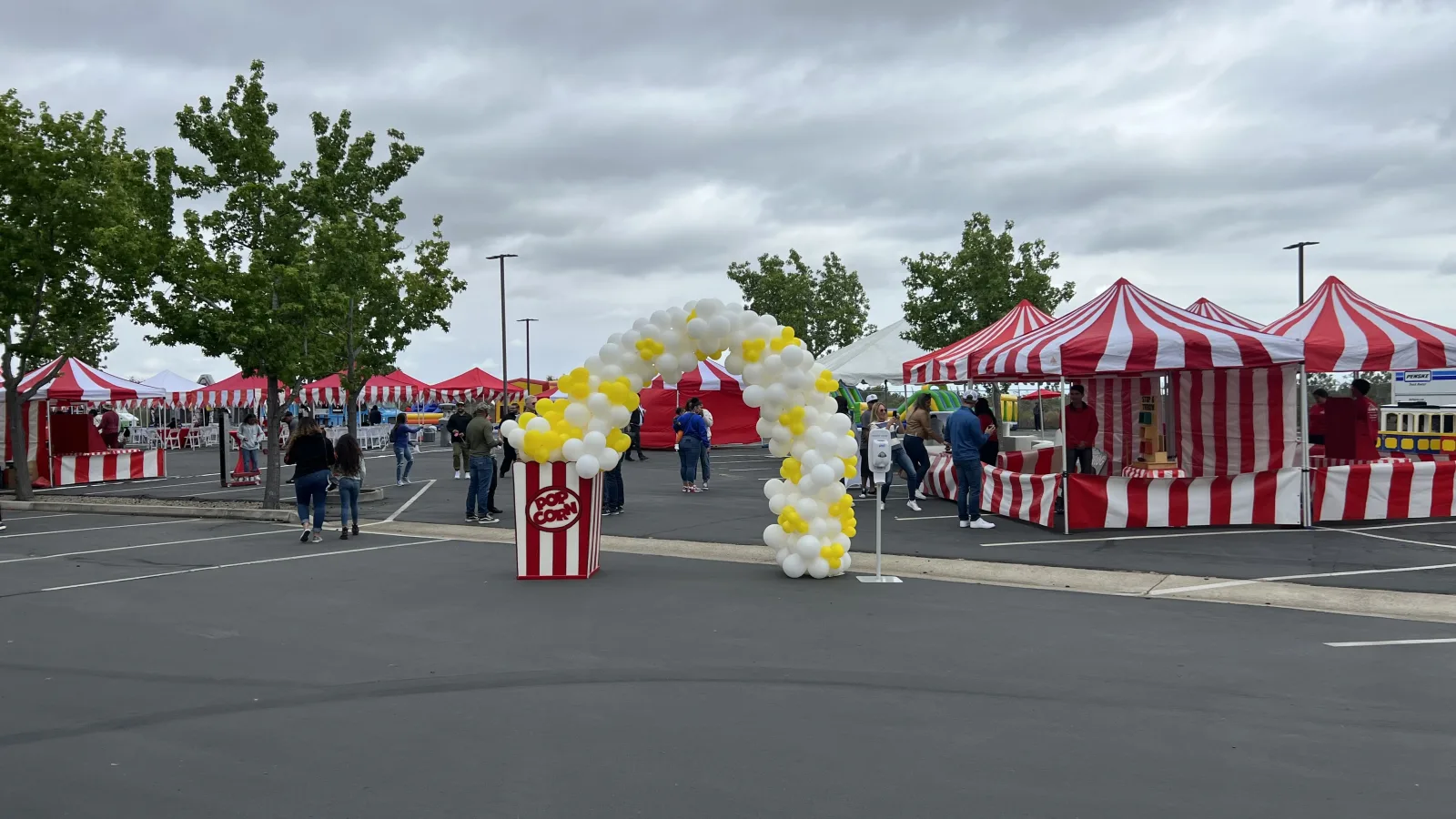 Red-and-white striped carnival booths set up across a school parking lot under an overcast sky, with a popcorn-themed balloon arch and a giant popcorn box prop at the entrance