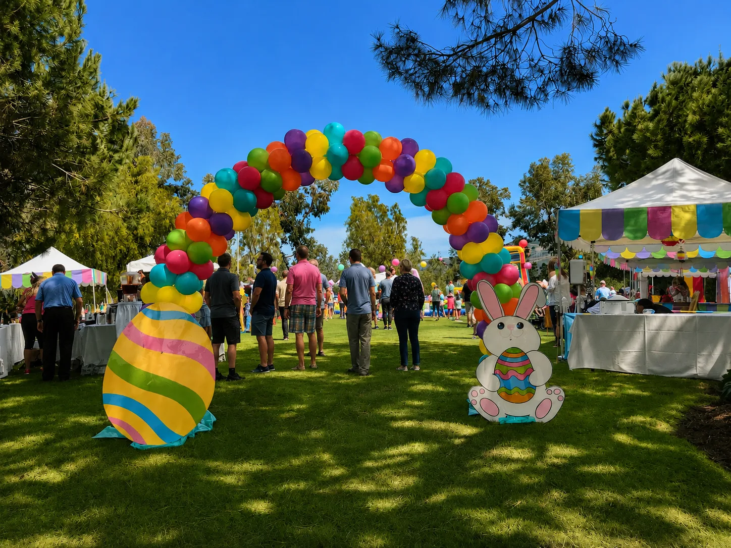 A rainbow balloon arch on grass at an Easter event, framed by a large decorated Easter egg cutout and a bunny cutout, with pastel-bunted booths and a crowd behind