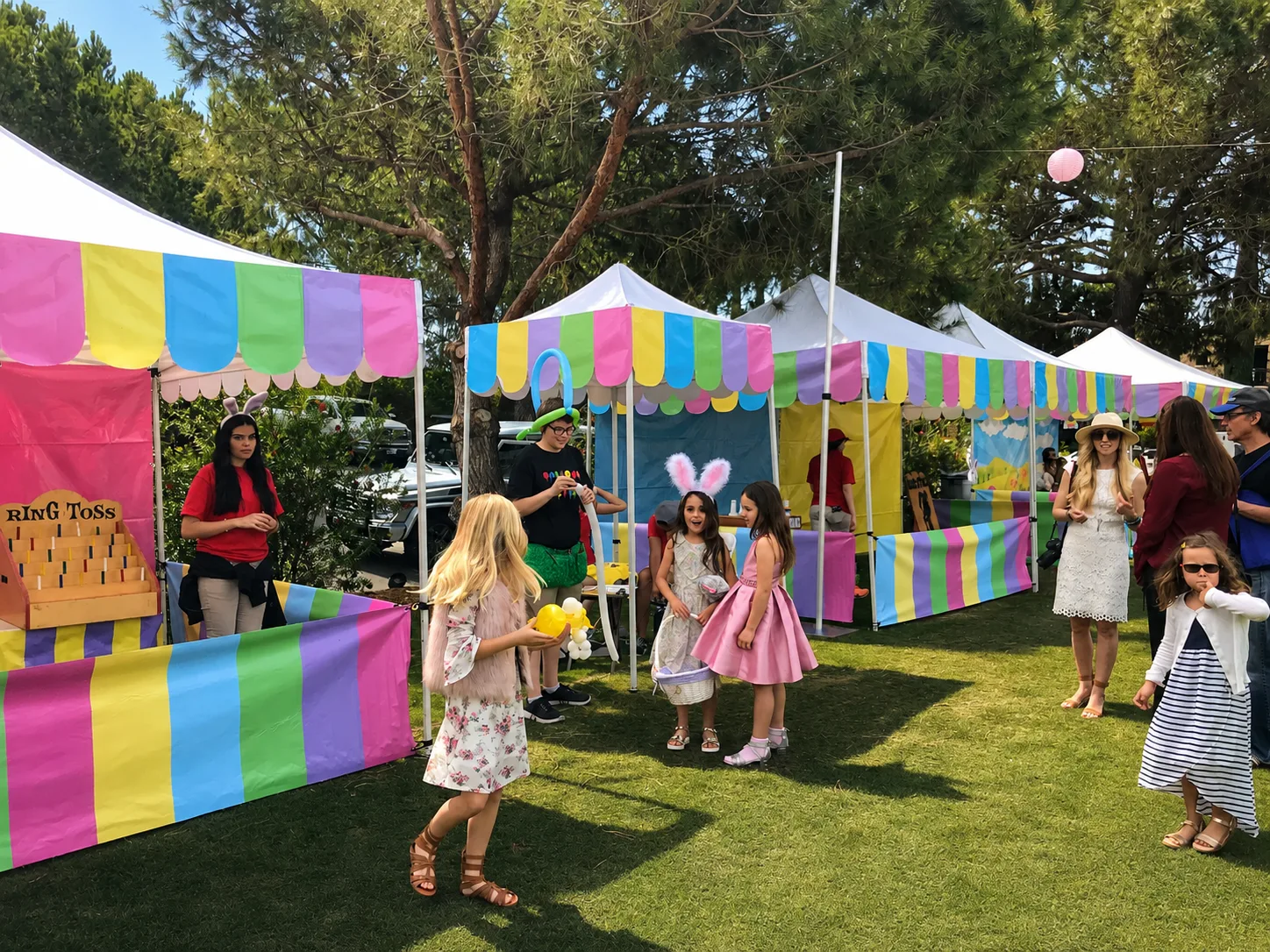 Pastel-striped carnival booths with scalloped valances at an outdoor Easter event, a wooden ring-toss game in the foreground, girls in bunny ears carrying Easter baskets, and a balloon twister at work