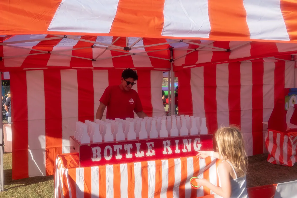 A child playing a Bottle Ring game at an orange-and-white striped carnival booth, one of the Halloween booth colors offered for Indio events