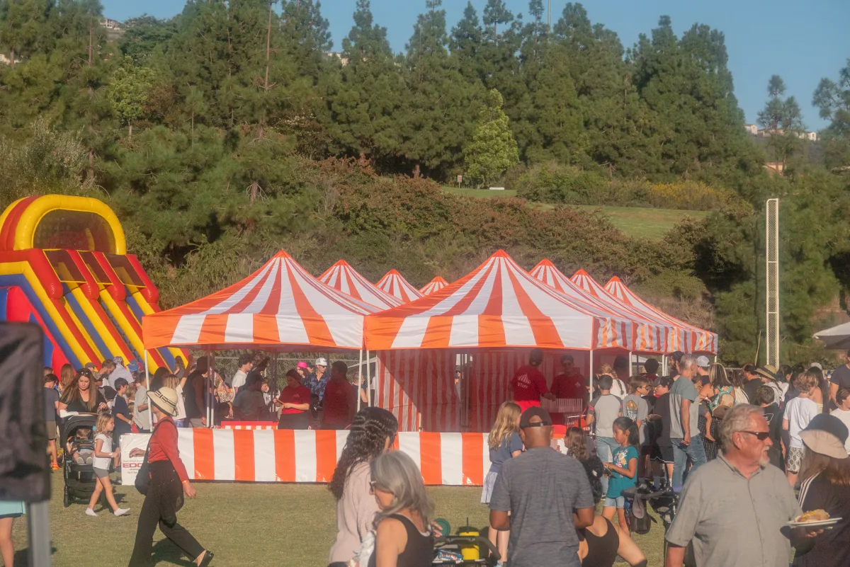 Rows of orange-and-white striped carnival booths on a grass field at a busy outdoor event, families queuing and an inflatable slide off to the side