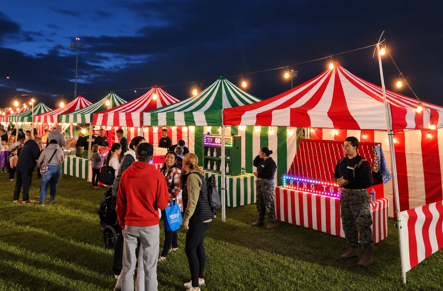 Red-and-white and green-and-white striped carnival booths under string lights at dusk, with a lit ring-toss marquee and a crowd gathered on the grass