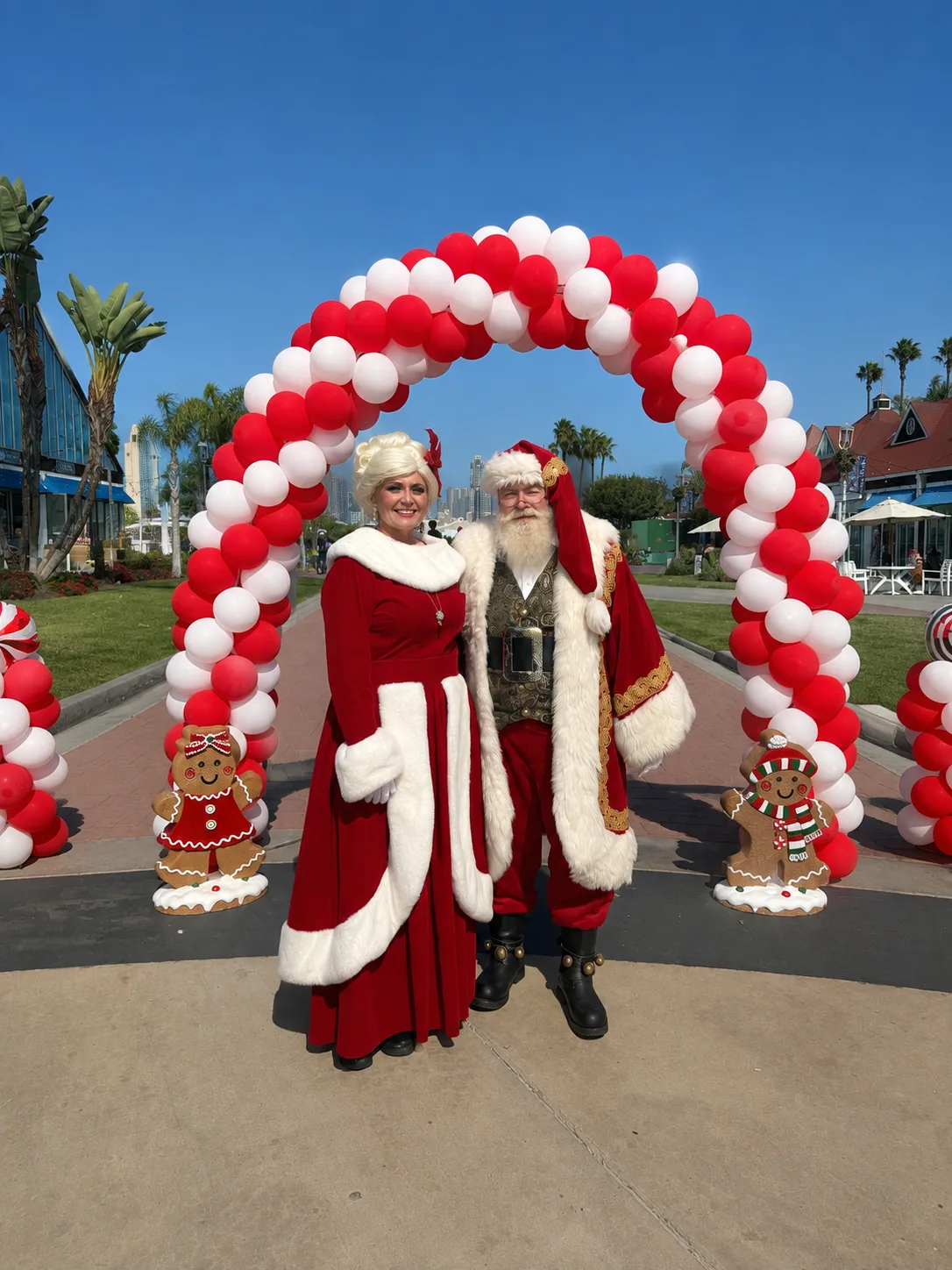 Santa and Mrs. Claus posing under a red-and-white balloon arch flanked by gingerbread figures at an outdoor holiday event